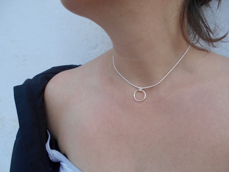 Pode incluir: Um colar tipo gargantilha de tom prateado com uma corrente fina e texturizada e um &uacute;nico pingente de anel circular liso centrado na frente. O colar repousa rente ao pesco&ccedil;o, exibindo um design met&aacute;lico simples com acabamento polido.
