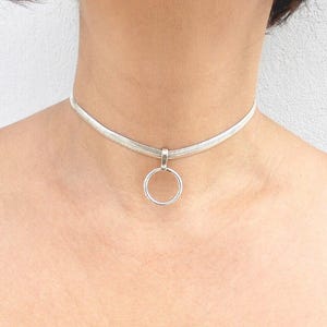 May include: A silver-toned choker necklace featuring a flat, woven herringbone chain design. The necklace includes a central circular pendant ring attached to the chain by a small, simple silver-toned bail, resting against the base of the neck.
