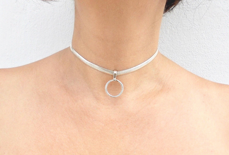 May include: A silver-toned choker necklace featuring a flat, woven herringbone chain design. The necklace includes a central circular pendant ring attached to the chain by a small, simple silver-toned bail, resting against the base of the neck.