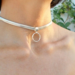 May include: A silver choker necklace with a herringbone pattern. The necklace has a small, round pendant hanging from the center. The jewelry is worn around the neck.