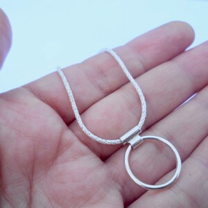 Pode incluir: Um colar de tom prateado com uma corrente fina de corda texturizada e um pingente circular liso preso por uma pequena argola retangular. O acabamento met&aacute;lico tem uma qualidade brilhante e reflexiva, com um design simples e uniforme.