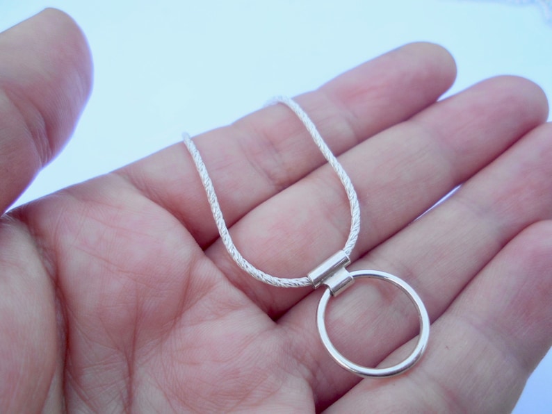 Pode incluir: Um colar de tom prateado com uma corrente fina de corda texturizada e um pingente circular liso preso por uma pequena argola retangular. O acabamento met&aacute;lico tem uma qualidade brilhante e reflexiva, com um design simples e uniforme.