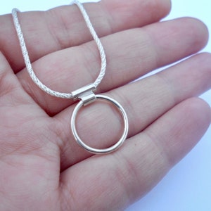 Pode incluir: Um colar de tom prateado com uma corrente fina e texturizada, ligada a um pendente circular liso. O pendente est&aacute; preso &agrave; corrente por uma pequena argola met&aacute;lica retangular, criando um design simples e limpo para esta pe&ccedil;a de joalharia.