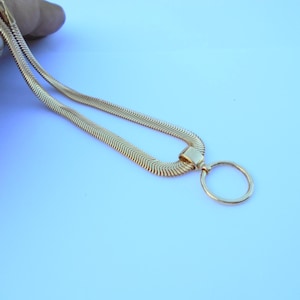 May include: A gold-colored necklace with a unique design. The necklace features a flat, herringbone chain and a circular pendant. The chain is held by a hand against a light blue background. The necklace is a stylish accessory.
