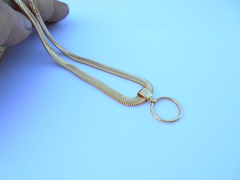 May include: A gold-colored necklace with a unique design. The necklace features a flat, herringbone chain and a circular pendant. The chain is held by a hand against a light blue background. The necklace is a stylish accessory.