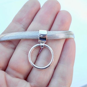 May include: A silver-toned bracelet with a herringbone pattern. A circular charm hangs from the bracelet. The bracelet is held in a hand against a light background.
