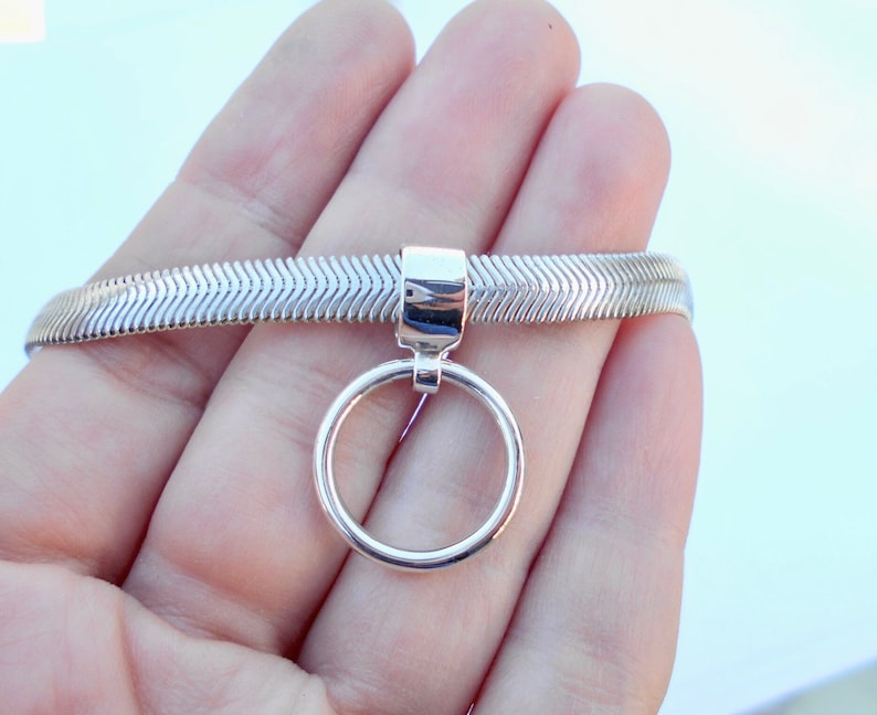 May include: A silver-toned bracelet with a herringbone pattern. A circular charm hangs from the bracelet. The bracelet is held in a hand against a light background.