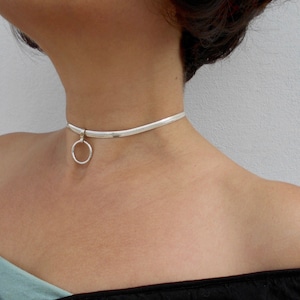 May include: A silver-toned snake chain choker necklace featuring a smooth, flexible band that sits closely around the neck. A single, circular silver-toned ring charm is attached to the center of the chain, hanging down slightly from the main band.