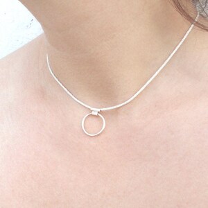 Pode incluir: Um colar tipo gargantilha de tom prateado com uma corrente fina e texturizada e um &uacute;nico pendente circular liso preso ao centro. O colar repousa sobre o pesco&ccedil;o, exibindo um design simples com um acabamento met&aacute;lico polido em toda a pe&ccedil;a.