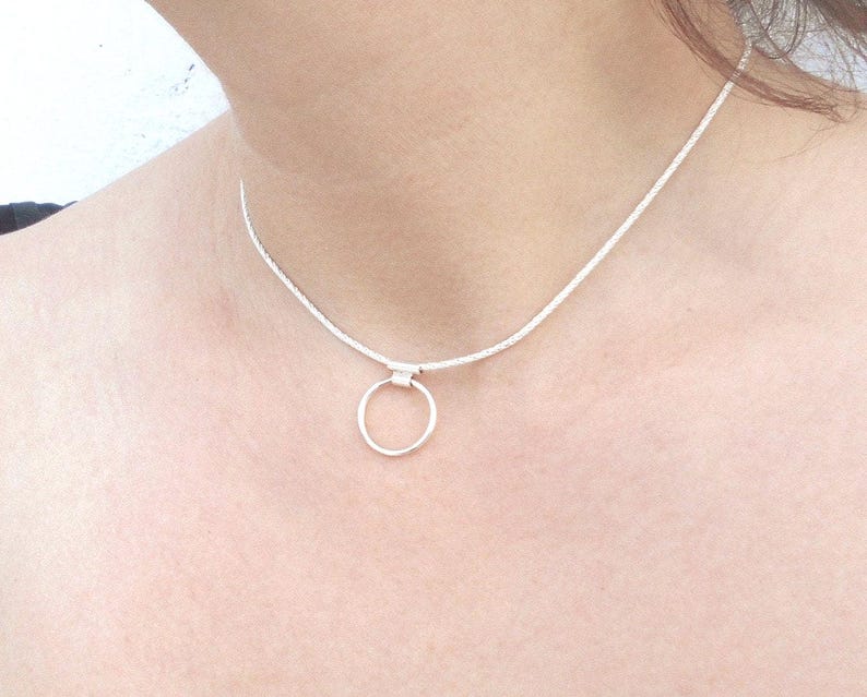 Pode incluir: Um colar tipo gargantilha de tom prateado com uma corrente fina e texturizada e um &uacute;nico pendente circular liso preso ao centro. O colar repousa sobre o pesco&ccedil;o, exibindo um design simples com um acabamento met&aacute;lico polido em toda a pe&ccedil;a.