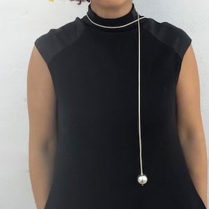 May include: A long silver chain necklace with a large silver ball pendant. The necklace is worn by a person wearing a black mock neck shirt.