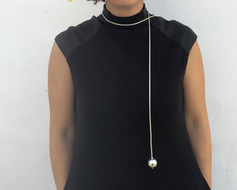 May include: A long silver chain necklace with a large silver ball pendant. The necklace is worn by a person wearing a black mock neck shirt.