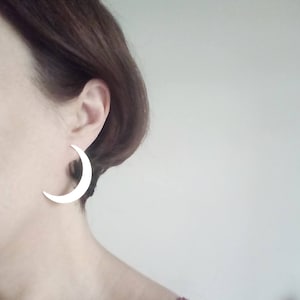 May include: A close-up of a person's ear wearing a large, white crescent moon earring. The earring is a simple, minimalist design, and the person has brown hair.