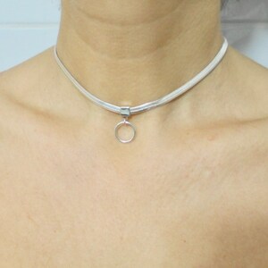 K&ouml;nnte beinhalten: Eine silberne Choker-Kette mit einem kleinen, runden Anh&auml;nger. Die Kette besteht aus einem d&uuml;nnen, flachen Metallband.