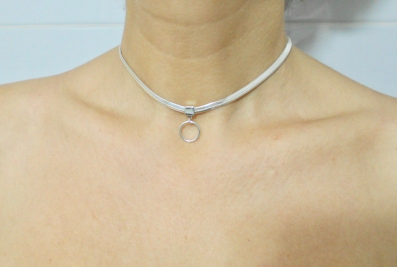 K&ouml;nnte beinhalten: Eine silberne Choker-Kette mit einem kleinen, runden Anh&auml;nger. Die Kette besteht aus einem d&uuml;nnen, flachen Metallband.
