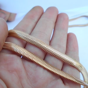 May include: A gold-colored herringbone chain necklace is held in a hand. The chain has a flat, flexible design with a repeating chevron pattern. The background is a plain white surface.