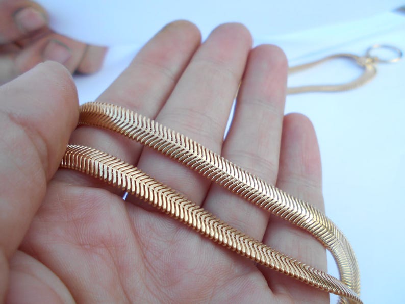 May include: A gold-colored herringbone chain necklace is held in a hand. The chain has a flat, flexible design with a repeating chevron pattern. The background is a plain white surface.