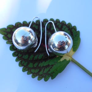 May include: A pair of silver dangle earrings with round, smooth, reflective balls. The earrings are hanging from a green leaf.