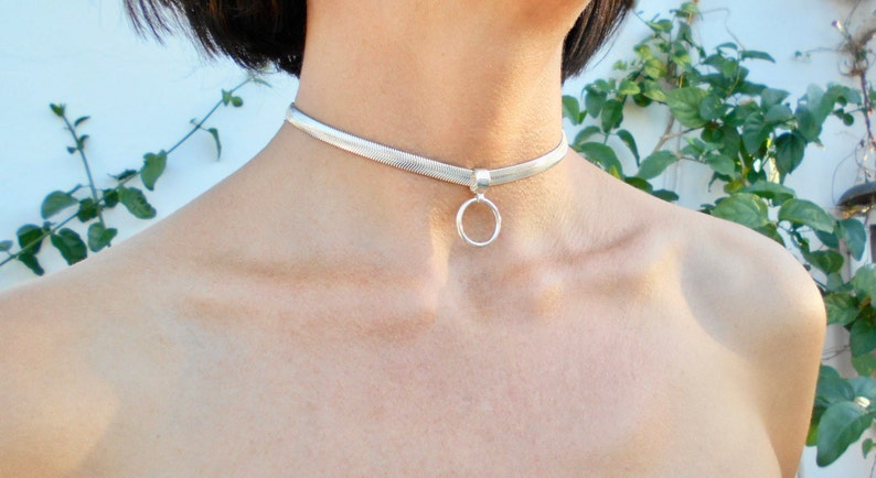 May include: A silver choker necklace with a herringbone pattern. The necklace features a small, round pendant. The jewelry is worn around the neck. The background includes green foliage and a white wall.