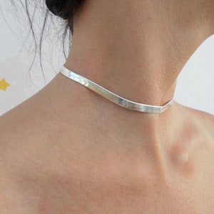 Pu&ograve; includere: Un girocollo in argento con un design piatto leggermente curvo. Il girocollo &egrave; indossato da una persona con i capelli tirati indietro.