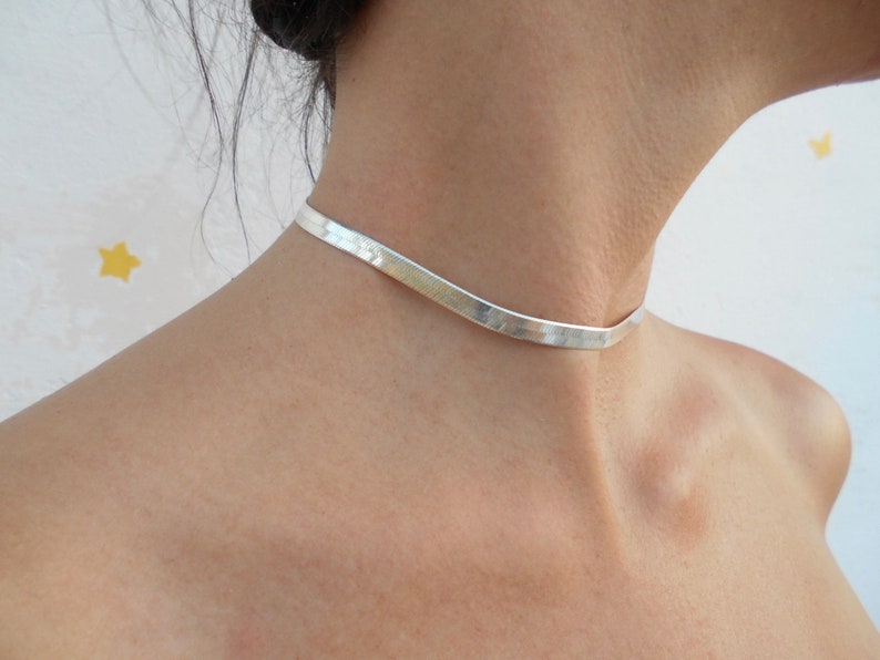 Pu&ograve; includere: Un girocollo in argento con un design piatto leggermente curvo. Il girocollo &egrave; indossato da una persona con i capelli tirati indietro.