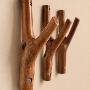 Puede incluir: Un conjunto de cuatro perchas de pared de madera, cada una con forma de rama natural. Las perchas son de color marrón cálido y están montadas en una pared beige claro. Se fijan con tornillos.