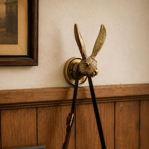 Puede incluir: Un gancho de pared color latón con forma de cabeza de conejo y orejas largas, montado sobre una base circular. Una cámara de estilo vintage plateada y negra con una correa de cuero marrón cuelga del gancho de conejo contra una pared de madera.