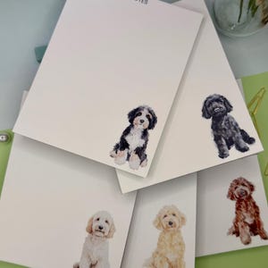 May include: A stack of white paper notepads with the word NOTES printed at the top. Each notepad features a watercolor illustration of a different dog breed, including a black and white dog, a black dog, a cream dog, a gold dog, and a brown dog.