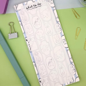 May include: A rectangular notepad with a white background and a pattern of dog illustrations in oval frames. The notepad has the text "shit to do" at the top. A light blue pen, a pink block, and gold paper clips are also visible.