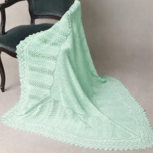 Pode incluir: Um cobertor de malha verde menta com um design detalhado em renda e uma borda ondulada. O cobertor está drapeado sobre uma cadeira verde escura, mostrando sua textura e padrão. A cor pastel suave sugere um item aconchegante e delicado.