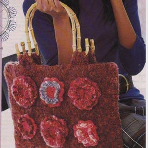 Pode incluir: Uma bolsa tote marrom avermelhada com alças de bambu e decorações florais. A bolsa apresenta seis apliques de flores vermelhas texturizadas, uma com um centro azul e cinza. As alças são feitas de bambu de cor clara.