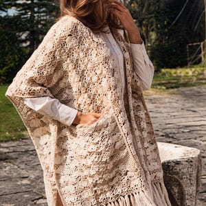 Pode incluir: Um poncho bege de crochê com detalhes de franjas. O poncho apresenta um padrão detalhado de trabalho aberto e dois bolsos frontais. A peça é usada sobre uma camisa branca abotoada e calças marrons. O poncho é de cor neutra.