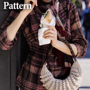 Pode incluir: Um PDF Pattern apresentando uma bolsa de crochê bege em formato de meia-lua com alça trançada escura. A bolsa é usada com uma blusa de manga comprida xadrez bordô e preta com cintura franzida e um lenço xadrez combinando ao redor do pescoço.