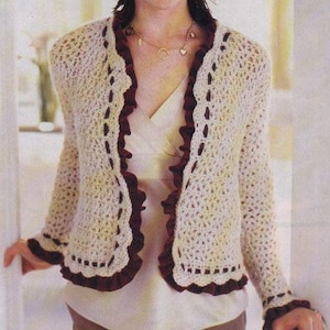May include: Cream-colored crochet cardigan with dark burgundy trim. The open-front sweater features a ruffled edge and is worn over a white top. The sleeves have a matching burgundy trim at the cuffs. The cardigan is a delicate, handmade garment.