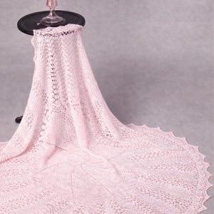 Pode incluir: Um delicado xale rosa claro com um intrincado padrão de renda, drapeado sobre uma pequena mesa preta. Uma taça de vidro rosa está sobre a mesa. A borda ondulada do xale se abre, criando um efeito suave e fluido.