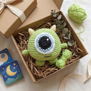 May include: A handmade green crochet monster toy with a large blue and black eye, presented in a brown gift box with paper filler. The box is tied with a cream ribbon. A ball of green yarn is also included.