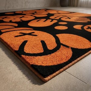 Pu&ograve; includere: Un tappeto rettangolare con un motivo ripetuto di volti arancioni in stile cartone animato con occhi a forma di X e mani arrotondate su sfondo nero. Il tappeto presenta una superficie testurizzata e trapuntata con un bordo arancione cucito.