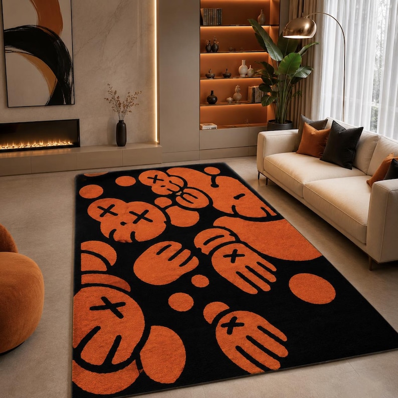 Pu&ograve; includere: Un tappeto rettangolare con un motivo ripetuto di teste e mani in stile cartone animato arancione su uno sfondo nero solido. Ogni disegno di testa e mano include un simbolo a croce distintivo per gli occhi, creando un audace motivo grafico.