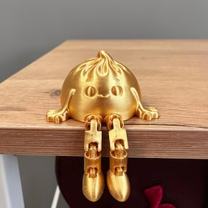 May include: A gold-coloured, 3D-printed figurine shaped like a dumpling with a pleated top, a smiling face, and two outstretched arms. The figure features two articulated, dangling legs with rounded feet that hang over the edge of a surface.