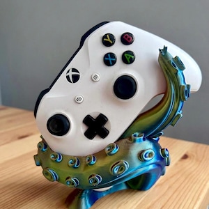 May include: A white Xbox controller sits on a 3D-printed stand. The stand is designed to look like an octopus, with blue, green, and gold tentacles. The controller has black buttons and coloured action buttons.