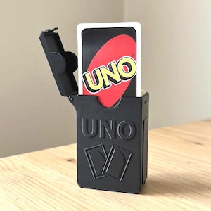 May include: Black plastic UNO card holder with a hinged lid, holding a single UNO card. The card has a black background with a red oval and the word "UNO" in yellow. The holder is on a light wood surface.