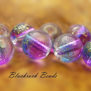 May include: A collection of clear, iridescent glass beads with a purple, blue, and gold shimmer. The beads are round with a hole for stringing. The text "Blackrock Beads" is written in cursive at the bottom.