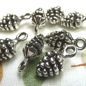 May include: Close-up of several small, silver-colored pinecone charms. Each charm has a detailed, textured surface and a loop for attaching to jewelry or other crafts. The charms are scattered on a patterned surface.