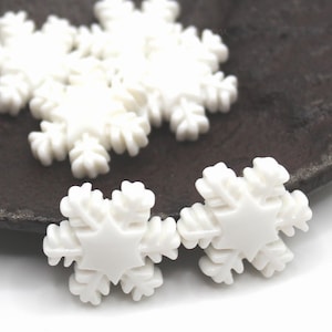 May include: White snowflake-shaped embellishments. The snowflakes have a star-shaped center and detailed, textured edges. Several are scattered on a dark surface, with two in the foreground. These decorative elements are ideal for crafts and seasonal projects.