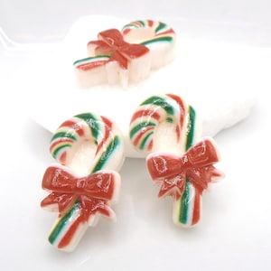 May include: Candy cane-shaped Christmas ornaments with red bows. The candy canes have red, green, and white stripes. The ornaments are on a white surface.