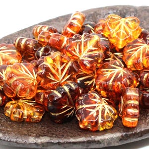 May include: A collection of amber-colored glass beads in various shapes, including maple leaf designs and cylindrical shapes. The beads have gold-colored accents and are displayed on a dark, textured surface.