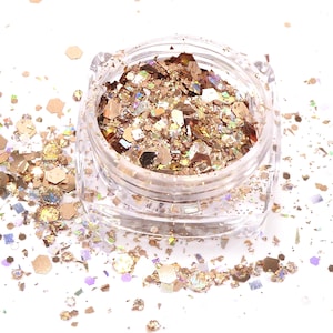 May include: A clear plastic jar filled with metallic gold and bronze cosmetic glitter flakes. The container is square-shaped with rounded corners, and loose, multi-sized hexagonal and irregular glitter pieces are scattered around the base.