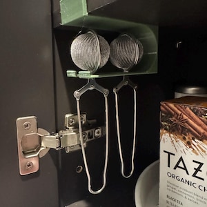 May include: Two stainless steel tea infusers hanging from a green holder inside a dark cabinet. The infusers have a mesh ball and long handles. A box of TAZO Organic Chai tea is visible.