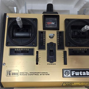 Könnte beinhalten: Ein goldfarbener Futaba FG Series digitaler proportionaler Funkfernsteuerungssender. Das Gerät verfügt über zwei silberne Joysticks, eine zentrale analoge Batteriestandsanzeige, mehrere schwarze Trimmregler, einen Netzschalter und verschiedene Kippschalter.