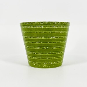 May include: A tapered, cylindrical plant pot in a vibrant grass green color. The exterior features a series of horizontal, raised ridges that wrap around the body, accented with a white, speckled, textured glaze finish.